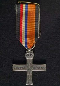 speldje van een kruis met een lintje er aan in de kleuren rood, wit, blauw en een bredere band oranje. 