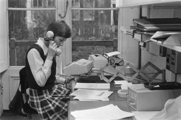 Oude zwart wit foto waarop een vrouw achter een typmachine een telefoon vasthoud waarmee ze belt.